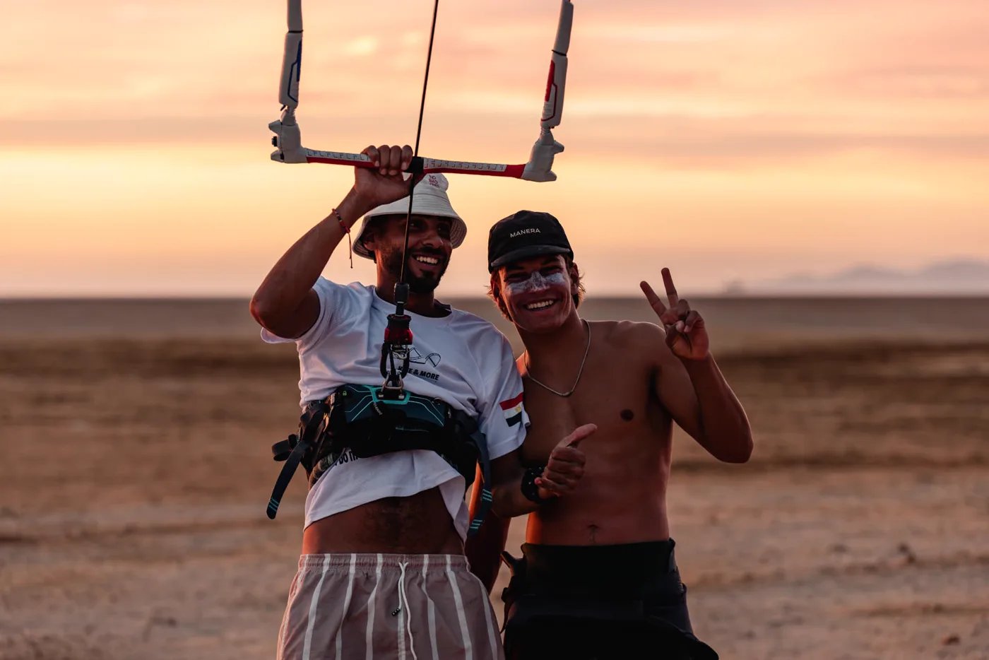 Two kitesurfers posing together on the beach at sunset, one holding the control bar overhead