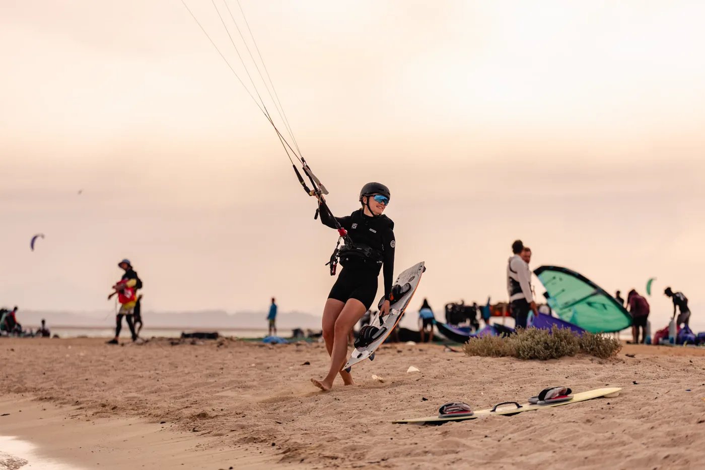 Woman in a helmet and wetsuit carrying her board and control lines across the beach at the camp