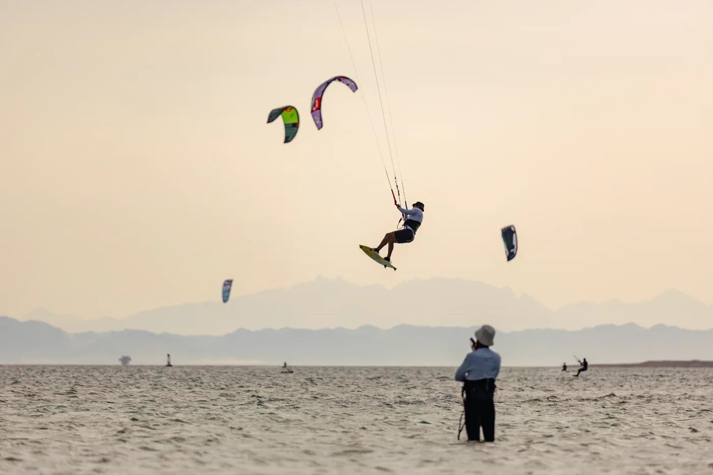 Kitesurfer mid-air over the water while a coach watches from the shallows