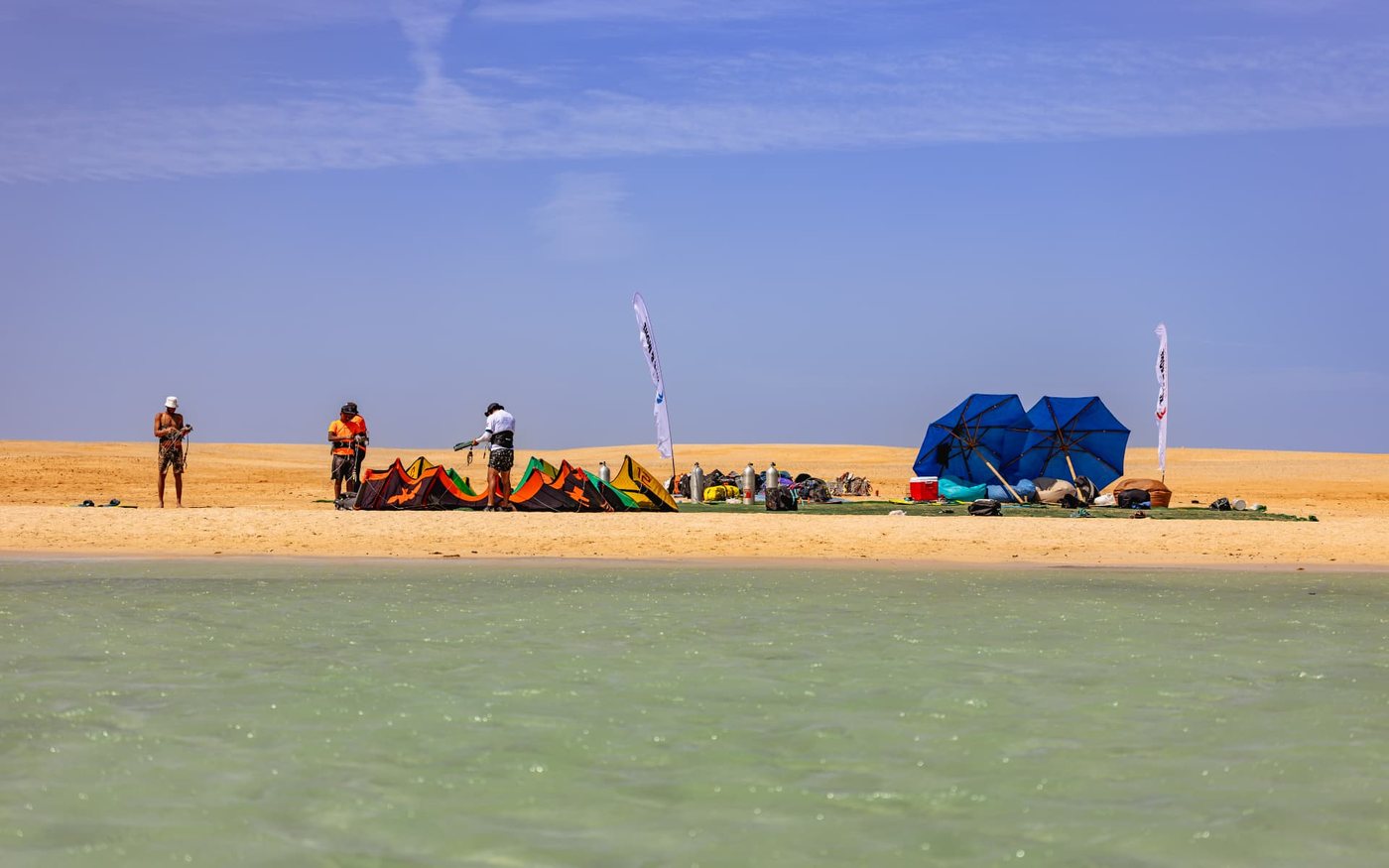 Kitesurfing gear being packed on a desert sandbar with blue umbrellas and turquoise water